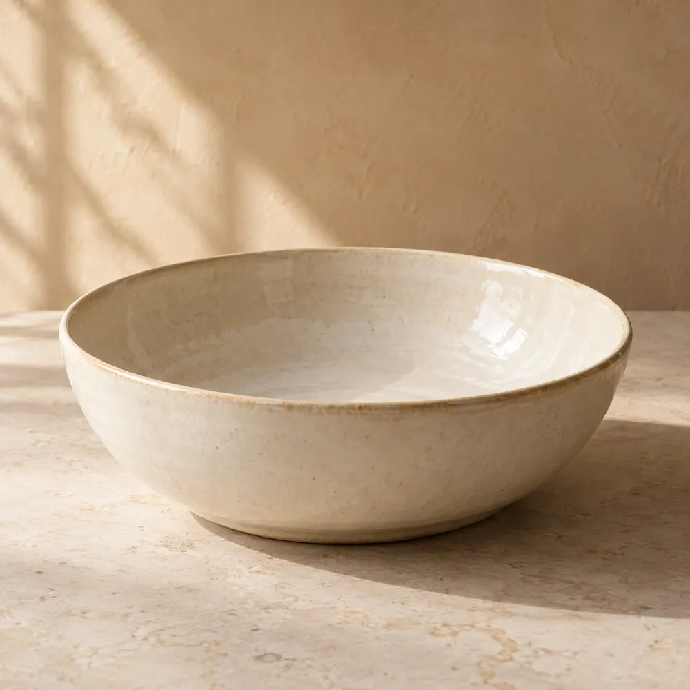A shallow warm ivory ceramic bowl on a sand-coloured surface
